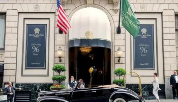 A vintage black convertible parked outside a grand building with flags (U.S. and green) and people nearby, classic city street scene.