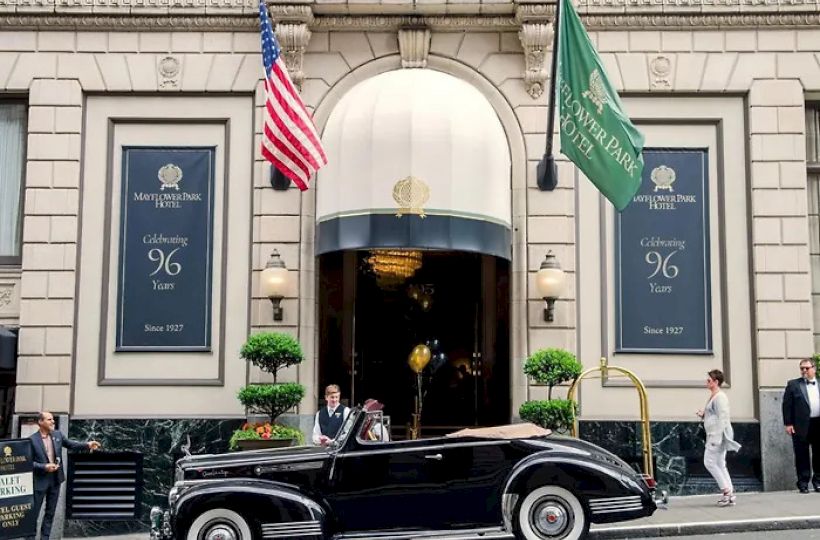 A vintage black convertible parked outside a grand building with flags (U.S. and green) and people nearby, classic city street scene.
