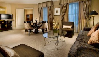 A spacious hotel suite with a seating area, glass coffee table, floral couch, draped windows, and a round dining table in the background.