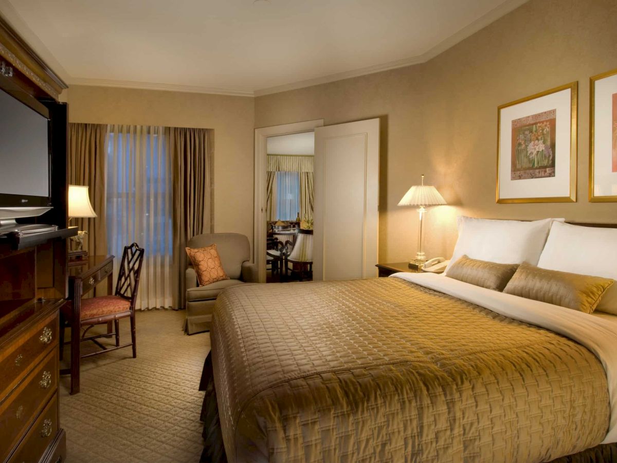 A hotel room with a large bed, matching brown bedding, nightstands, lamps, a desk and chair, a TV cabinet, and warm beige walls with framed art. The area looks neat and cozy.