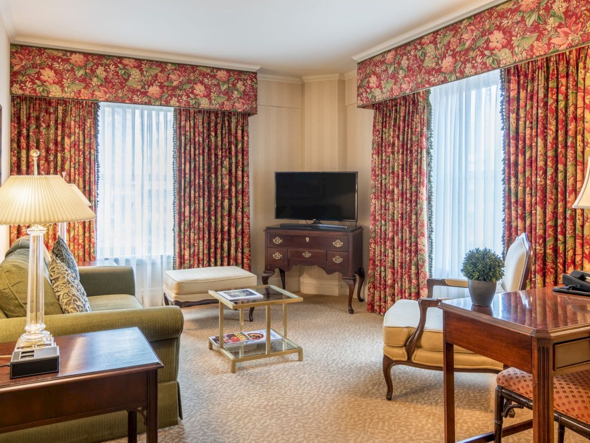 A traditional hotel suite with floral drapery, a green sofa, wooden furniture, a TV on a dresser, a glass coffee table, and a desk with a lamp and chair.