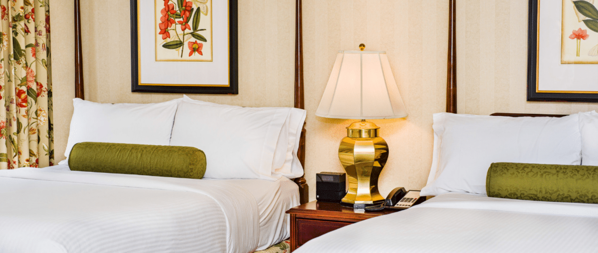 Two neatly made hotel beds with white linens and green bolster pillows, a bedside lamp, framed botanical art, and a wooden nightstand between them.