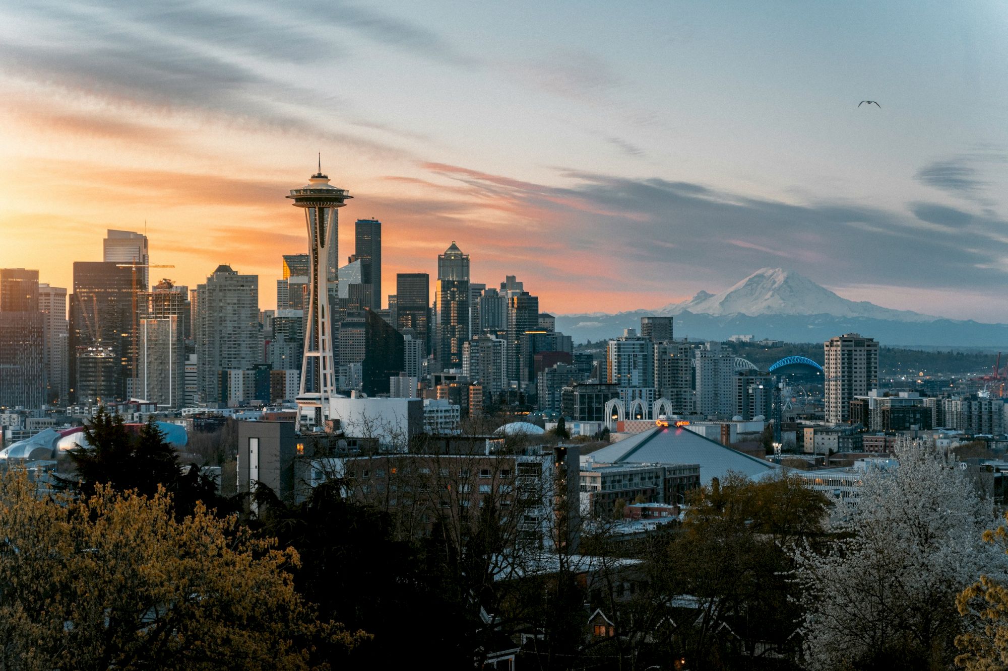 Seattle skyline at sunset with Space Needle, tall buildings, and distant mountains; warm orange sky fades to blue, trees in foreground.