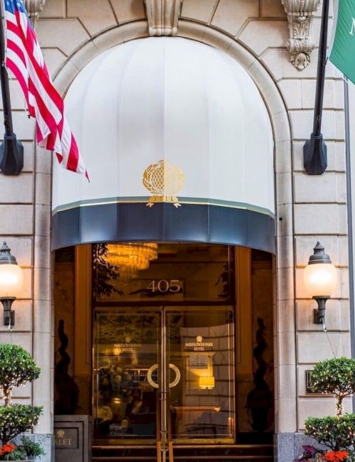 A grand hotel entrance with two banners celebrating 95 years, flags (American and green flag), ornate doors, and potted plants flanking the doorway. ending.