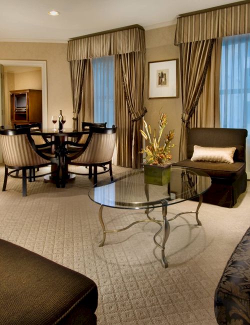 A luxurious hotel suite with a seating area, glass coffee table, patterned sofa, armchairs, dining table, curtains, lamps, and a flat-screen TV in the corner.