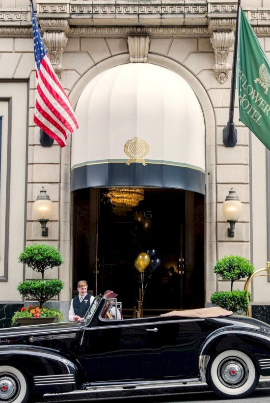 A classic black vintage car sits in front of the hotel entrance, with flags, doormen, and banners reading &ldquo;97&rdquo; beside the doors.