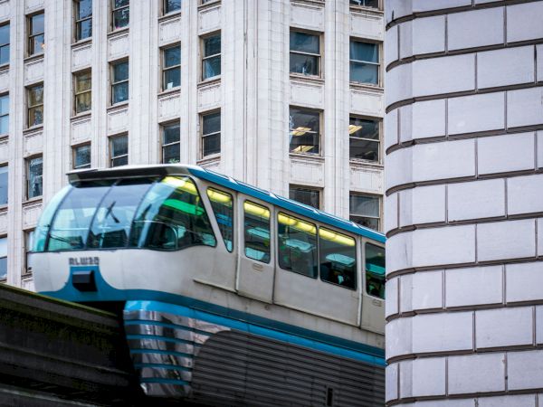 A futuristic monorail car peeks between tall urban buildings, riding along an elevated track beside a white brick wall.