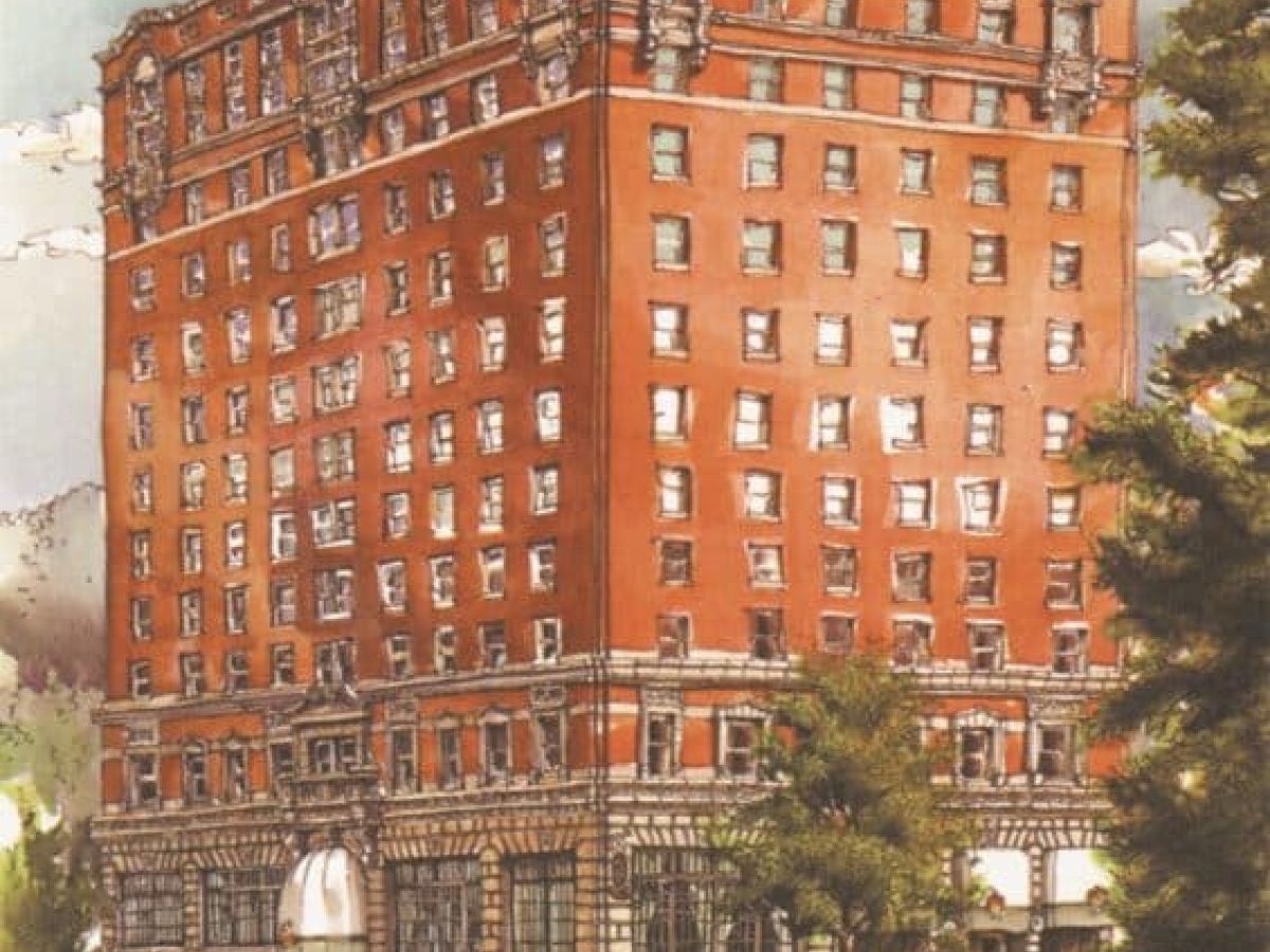 A tall red brick building with many windows sits on a bustling street, with storefronts on the ground floor and trees nearby.