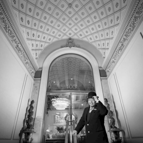 A black-and-white photo of a grand classical hallway or cathedral-like archway, featuring ornate columns, a vaulted ceiling, and a person walking toward a lighted doorway.