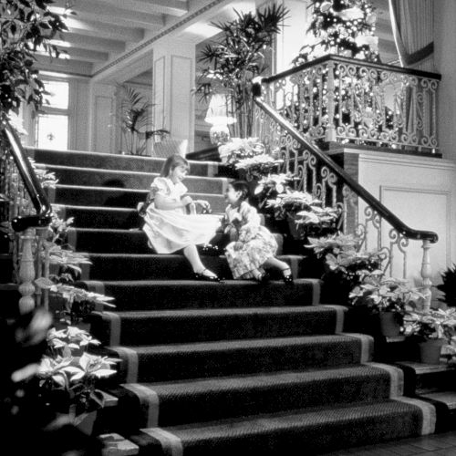 Two young girls in white dresses sit on a grand stairway lined with festive florals and a decorated railing, chatting beneath a tall, ornate Christmas tree.