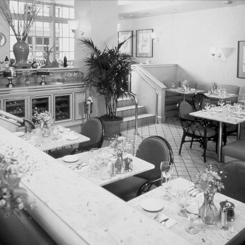 A black-and-white office or classroom scene with people seated around desks, papers scattered, a wall clock, and framed art in the background.