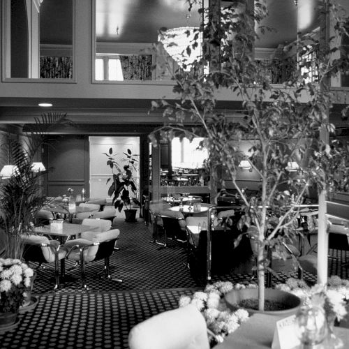 A black-and-white street scene with an elderly man sitting by a doorway, a bicycle, and shopfronts framed by a narrow alley.