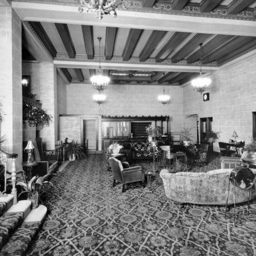 A vintage-style living room or studio with a long table, scattered chairs, antique d&eacute;cor, and a patterned carpet under string lights.