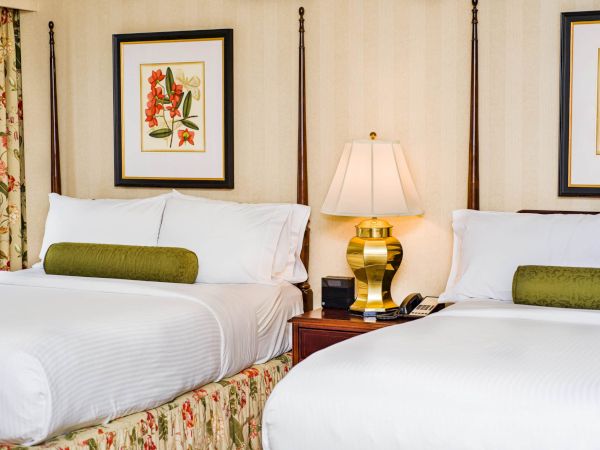 Two tidy double beds in a floral-lit hotel room with framed art, a bedside lamp, and green bolster pillows, neatly set.