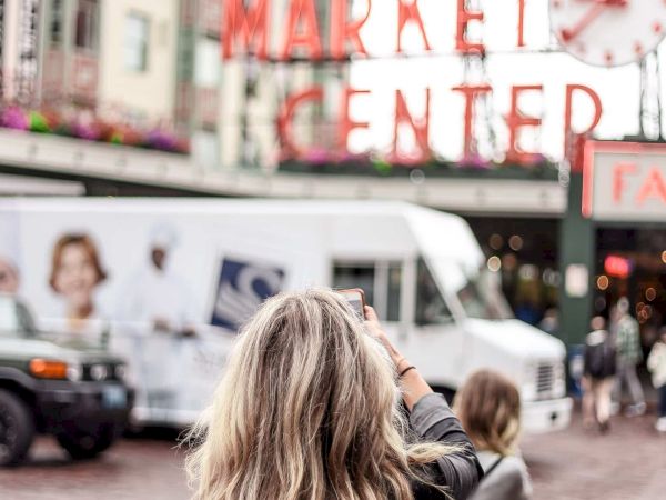 A woman with a backpack is taking a photo of a public market sign in a busy street. (140 characters)