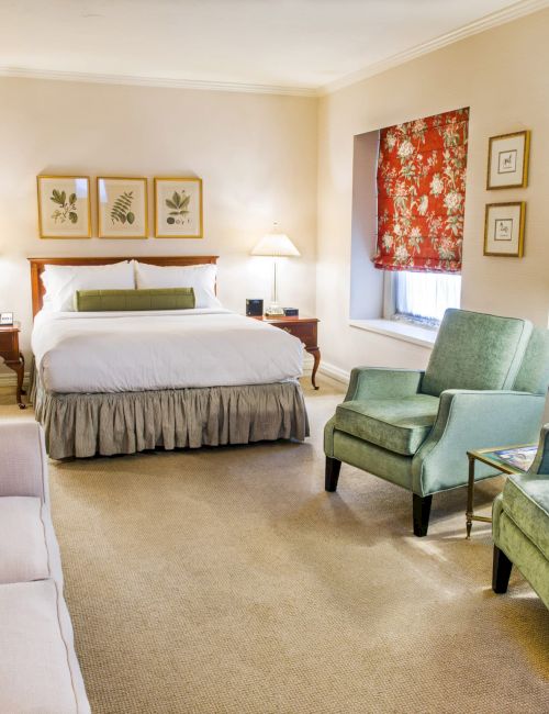A cozy hotel suite with a king bed, two nightstands and lamps, a beige sofa, two green chairs, patterned red window valances, and soft lighting.