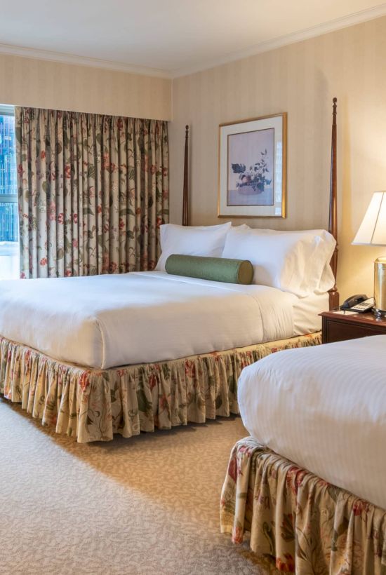 Two double beds in a bright hotel room with floral bedskirts, a seating armchair, nightstand lamps, and sheer curtains by a window.