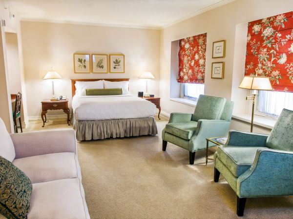 A deluxe hotel suite with a king bed, two green armchairs, a sofa, lamps, and floral red window coverings, creating a cozy, classic room.