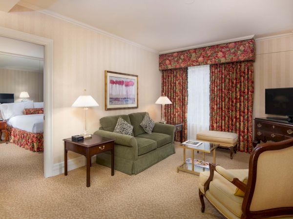 A hotel suite living area with a green sofa, side tables, lamps, a coffee table, patterned drapes, a TV on a dresser, and an adjacent bedroom visible through a doorway.