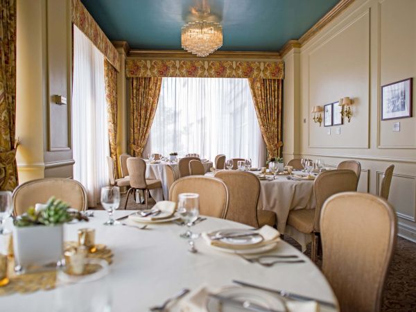 A fancy dining room set for a formal meal: round tables with white linens, gold chairs, chandeliers, paneled walls, and large windows with drapes.