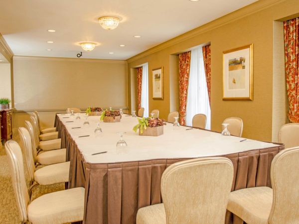 A long U-shaped table setup in a hotel conference room with beige chairs, draped curtains, and wall art, ready for a meeting.