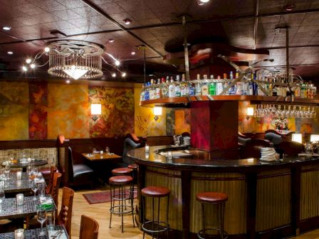 A cozy bar with a curved wooden counter, hanging lights, shelves of bottles, and booths in warm, colorful decor, ready for patrons.