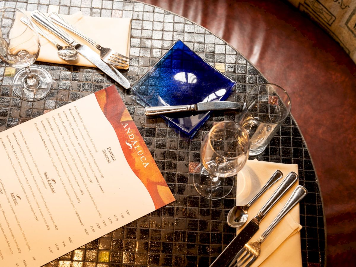 A round table set for dining with a menu, glasses, silverware, a blue ashtray, and wine on a mosaic surface.