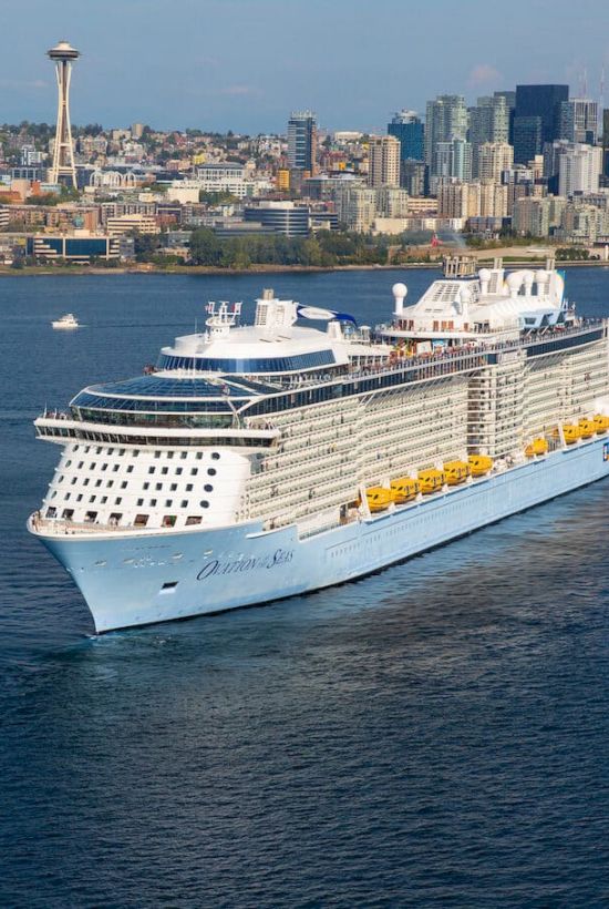 A large white cruise ship sails near a city waterfront, with tall buildings in the background and calm blue water. Top it at 140 characters, always ending the sentence.