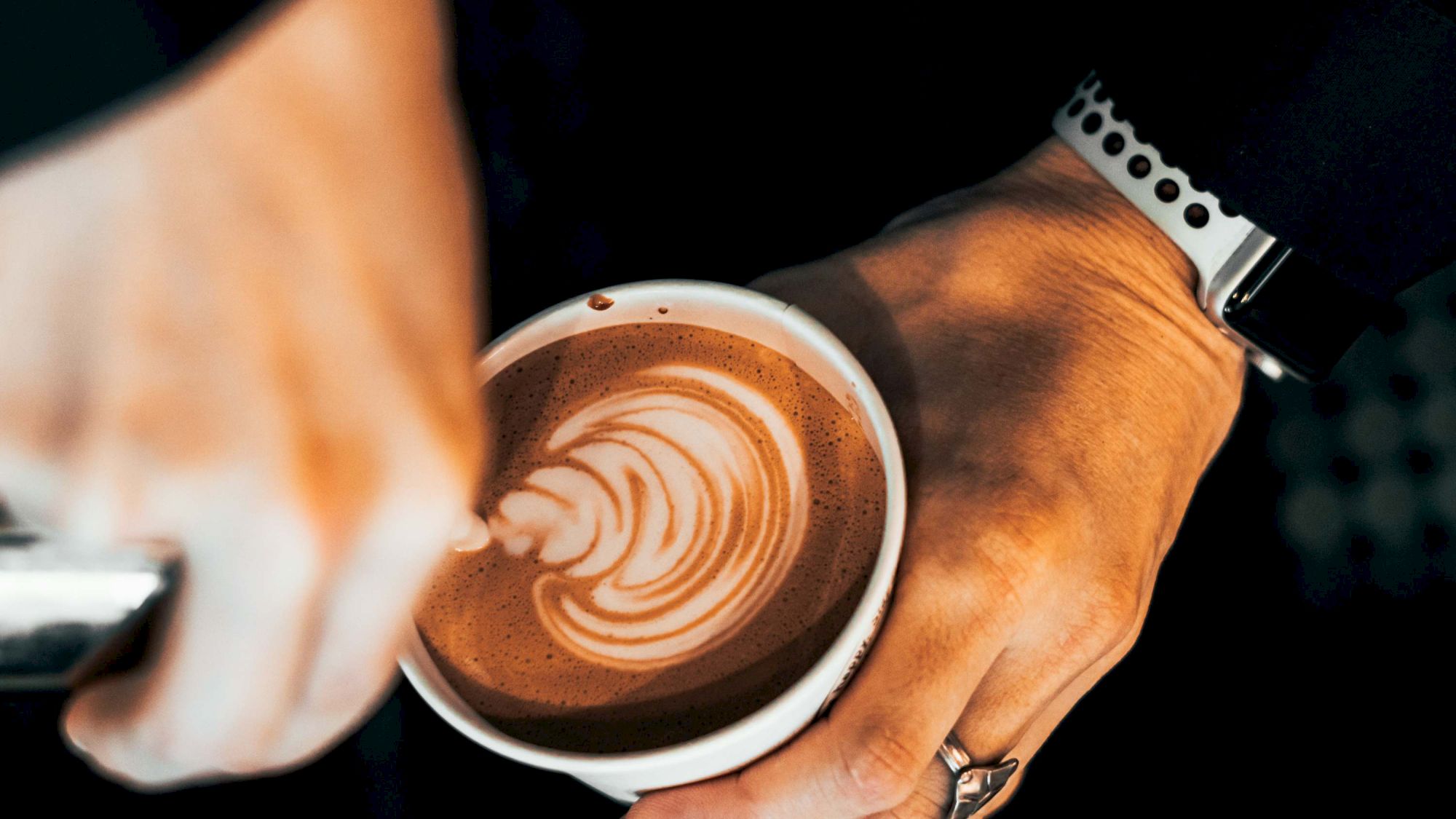 A barista pours latte art into a cup, swirling white foam into a rosette pattern as a hand grips the cup and a wristwatch and ring appear, framing the shot.