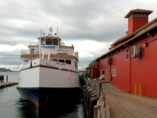 A large white boat named &ldquo;Nargosy&rdquo; docked at a wooden pier beside red waterfront buildings; cloudy skies loom overhead.