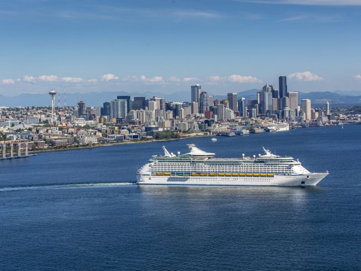 A large white cruise ship sailing past a city skyline along a blue harbor, with mountains in the distance. Top it at 140 characters, always ending the sentence.