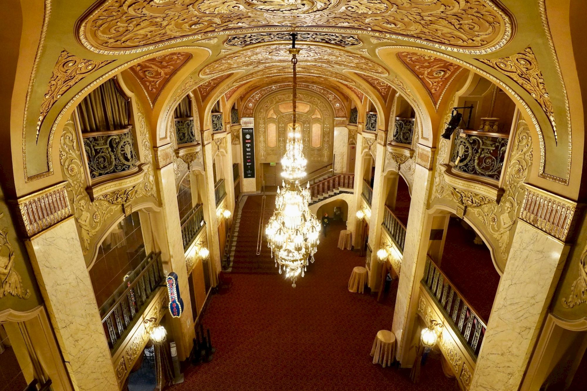 Opulent theater lobby with gilded arches, balconies, and a grand chandelier illuminating a red-carpeted floor, hinting at vintage elegance.