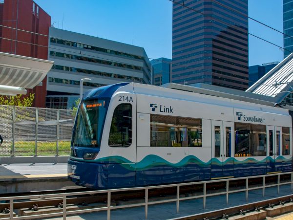 A blue and white Link light rail train at a city station, with modern buildings in the background and a pedestrian on the platform.