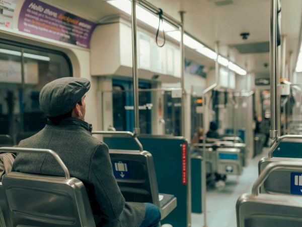 A man sits alone on a bus, back turned, wearing a dark coat and hat, looking ahead as the empty seats line the quiet interior.