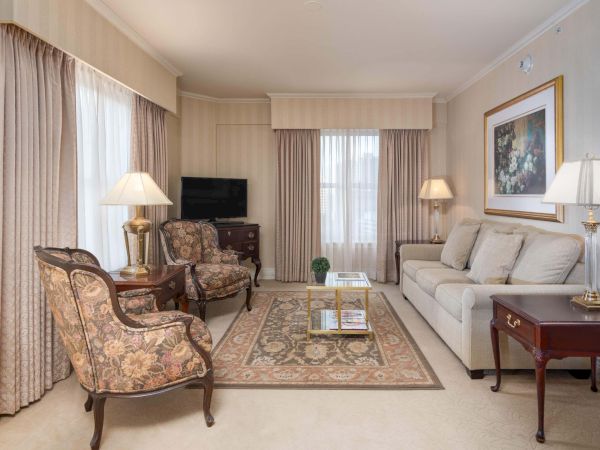 A classic, elegant living room with beige walls, cream sofa, patterned armchairs, wooden side tables, and a central coffee table on a traditional rug.