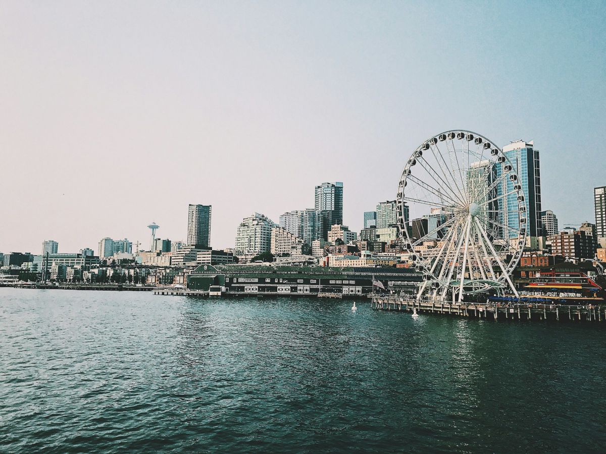 A city waterfront with a large Ferris wheel by the pier, modern buildings in the background, and calm water in the foreground.