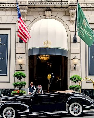 A vintage black car pulls up at a grand hotel entrance, flags flying, with uniformed staff and guests in front. There are blue banners and potted trees flanking the doorway.