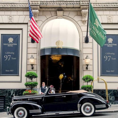 A small storefront with a rounded doorway, two large windows, a striped awning, potted plants, and a white van parked in front.