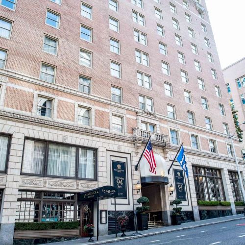 A busy urban street with mid-rise storefronts, pedestrians, and parked cars under clear weather, showing a mix of shops and office buildings.