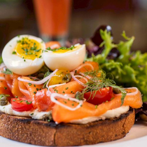 A slice of open-faced toast topped with smoked salmon, cream cheese, onions, capers, herbs, and a boiled egg, served with greens on the side.