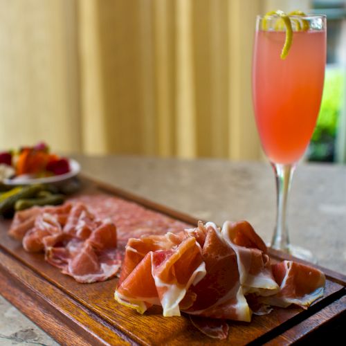 A wooden charcuterie board with assorted cured meats and an elegant pink cocktail on a marble counter, set by a bright window.