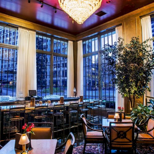 A stylish hotel lounge or restaurant with large windows, cozy seating, a bar, warm lighting, plants, and a chandelier overhead.