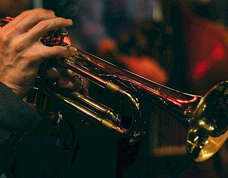 A close-up of a musician's hands playing a brass trumpet, dim stage lighting in the background, capturing a live jazz vibe.