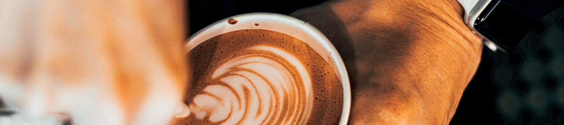 A barista pours latte art into a cup, creating a rosette design with a velvety foam, while the other hand holds the cup steady.