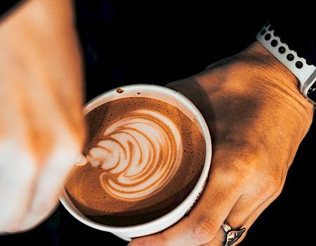 A barista pours latte art into a cup, creating a rosette design with a velvety foam, while the other hand holds the cup steady.