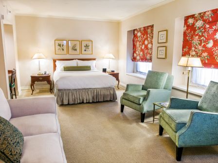 A cozy hotel room with a king bed, two green armchairs, a beige sofa, two lamps, red floral window coverings, and framed art on the walls.