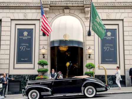 A luxurious black vintage car sits before a grand entrance of a hotel, with American and green flags, banners showing &ldquo;97&rdquo; and a doorman nearby, including people in formal attire.
