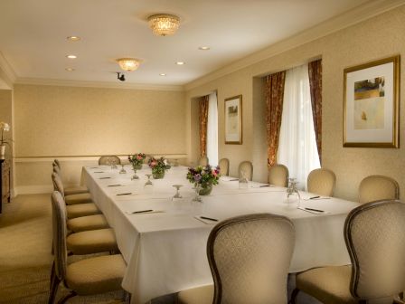 A formal dining room set for a meeting, with a long white-clothed table, floral centerpieces, and beige chairs along a sunlit wall.