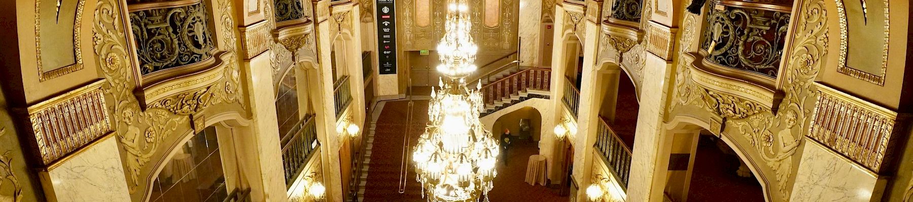 A grand, ornate theater lobby with gilded arches, balconies, and a large chandelier, viewed from above.