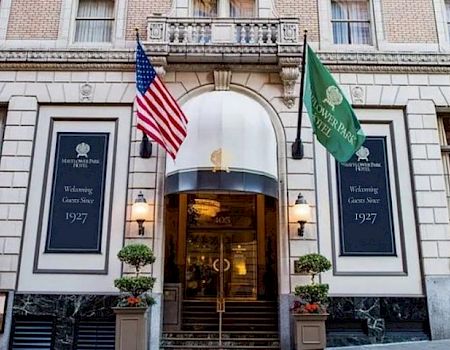 A grand hotel entrance with American and green flags, stone facade, and steps leading to a revolving door. (140 chars max, ending with a period)